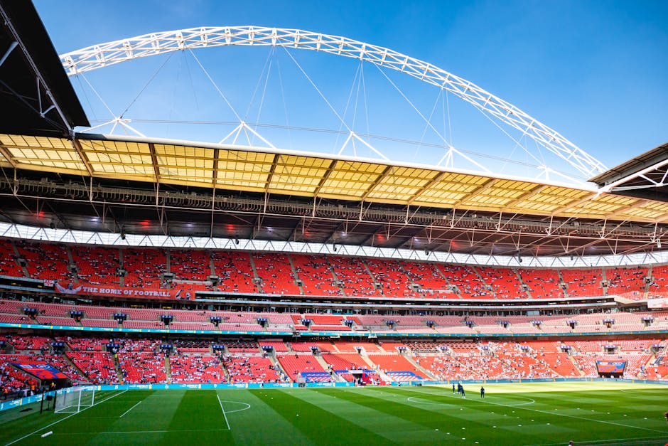 A large, modern stadium featuring a vibrant green soccer field with neatly mown grass and white boundary lines, surrounded by multiple tiers of red seating. The stadium roof, supported by metal framew