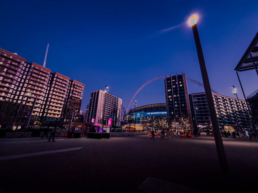 A nighttime cityscape featuring modern high-rise buildings with illuminated windows surrounding a large, circular Ferris wheel in the background, partially visible against a deep blue sky. The area in